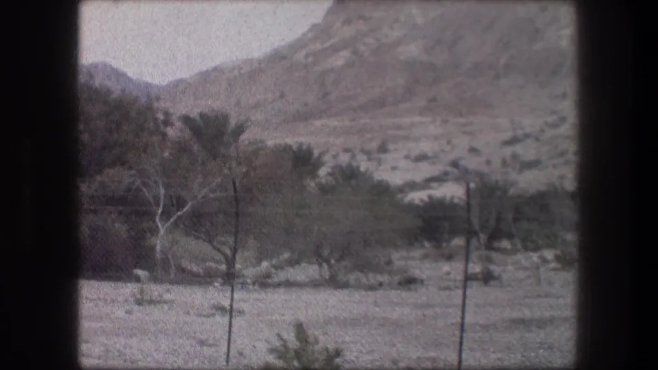 1976 Bethlehem 8mm frame with dry open ground, fence posts, palms, shrubs, and a rugged mountain slope.
