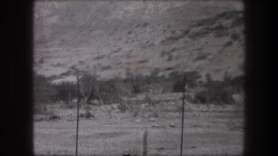 1976 Bethlehem-area home-movie frame with wire fencing, low brush and a pale rocky slope beyond the sandy foreground.