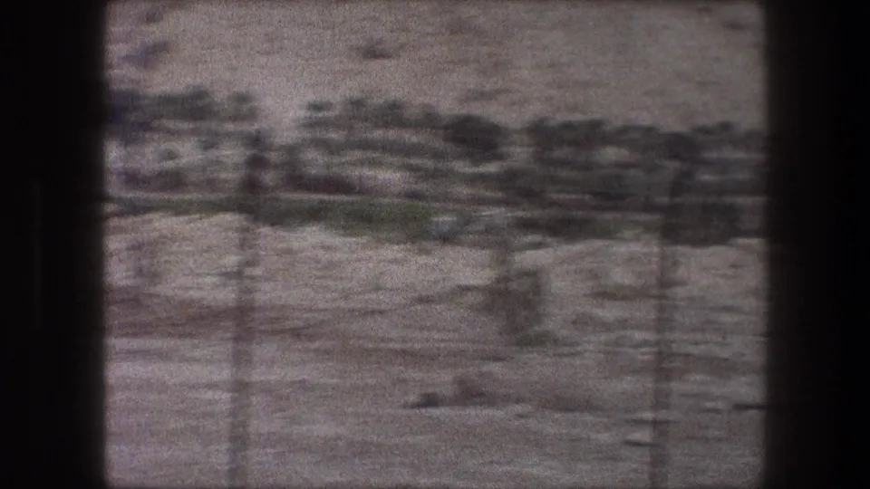 Handheld 8mm view of a dry Bethlehem-area landscape, with a blurred tree line and fence shadows or posts in front.