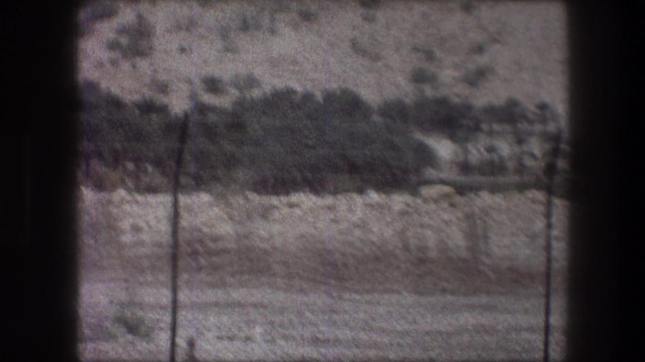 Barren rocky hillside seen through a dark window frame with sparse brush and fence posts.