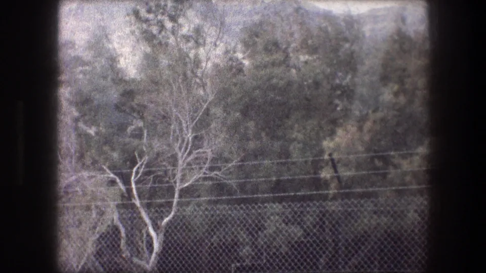 1976 8mm frame near Bethlehem with chain-link fencing, power lines, pale trees, and a rocky hillside beyond.