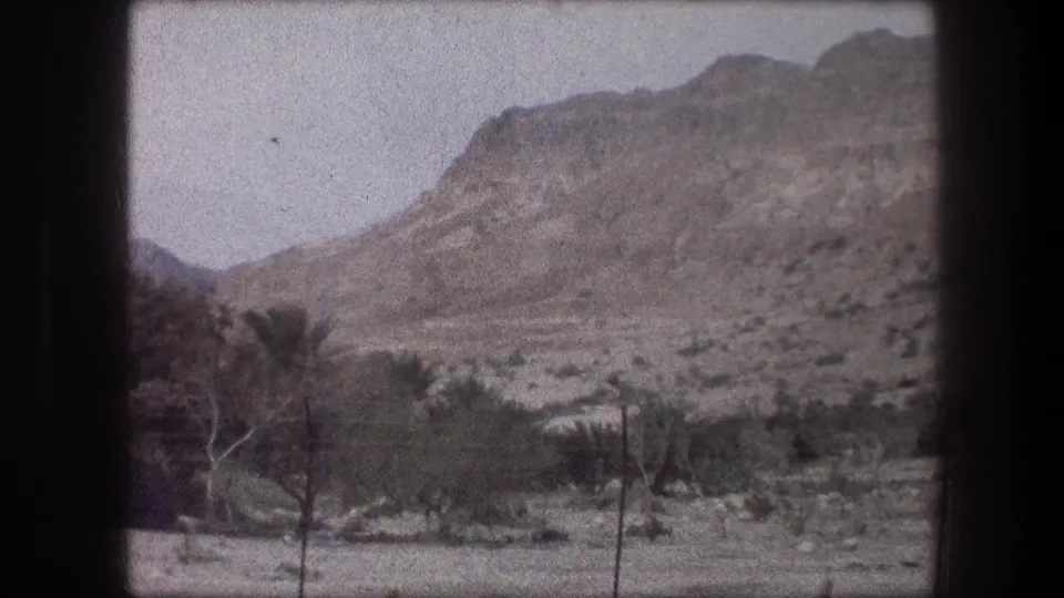 1976 8mm view near Bethlehem showing a rocky mountainside, sparse roadside trees, palms, and fence posts under a hazy sky.