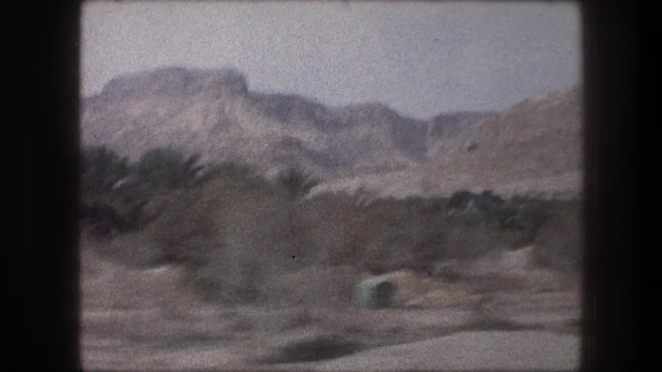 1976 8mm frame near Bethlehem with palm trees, dry roadside terrain, and distant rugged mountains.