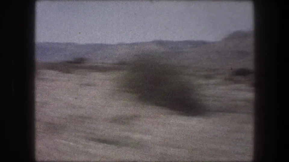 Moving 8mm frame from the Bethlehem area showing pale desert ground, a blurred shrub, and low hills on the horizon.