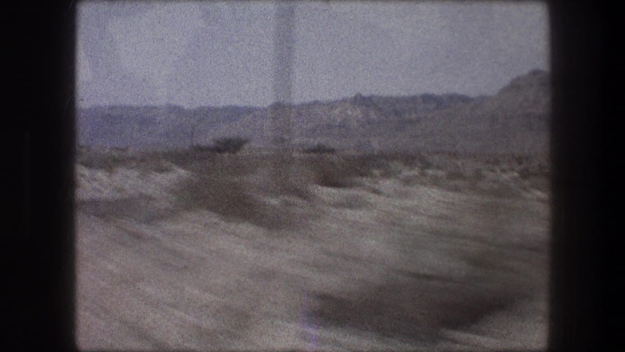 Arid roadside terrain with scrub and distant hills seen from a moving vehicle.