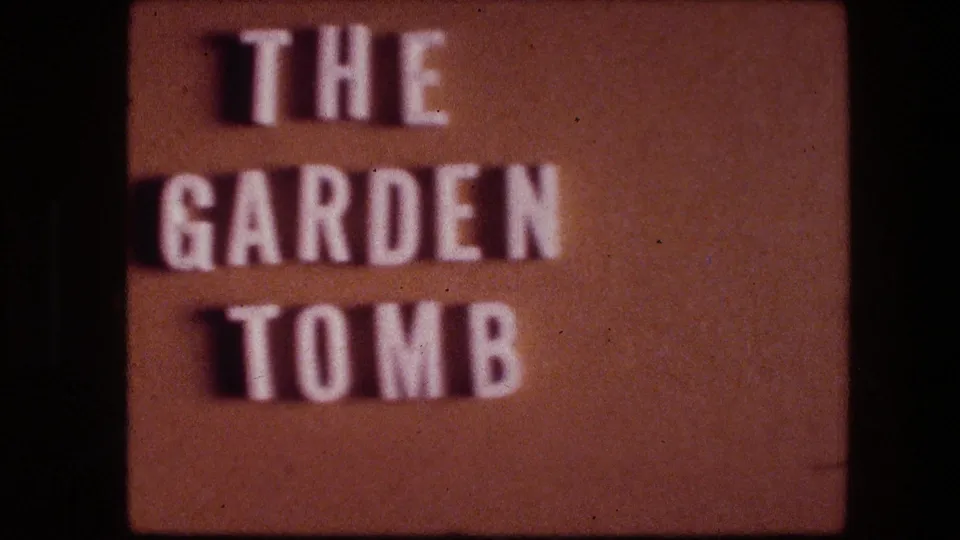 The Garden Tomb sign in Jerusalem appears in soft focus, with white raised lettering on a brown textured background.