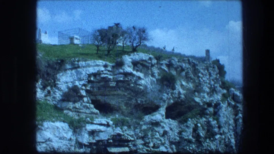 Garden Tomb area in Jerusalem, shown as a rocky cliff face below grass, scattered trees, and a clear blue sky.
