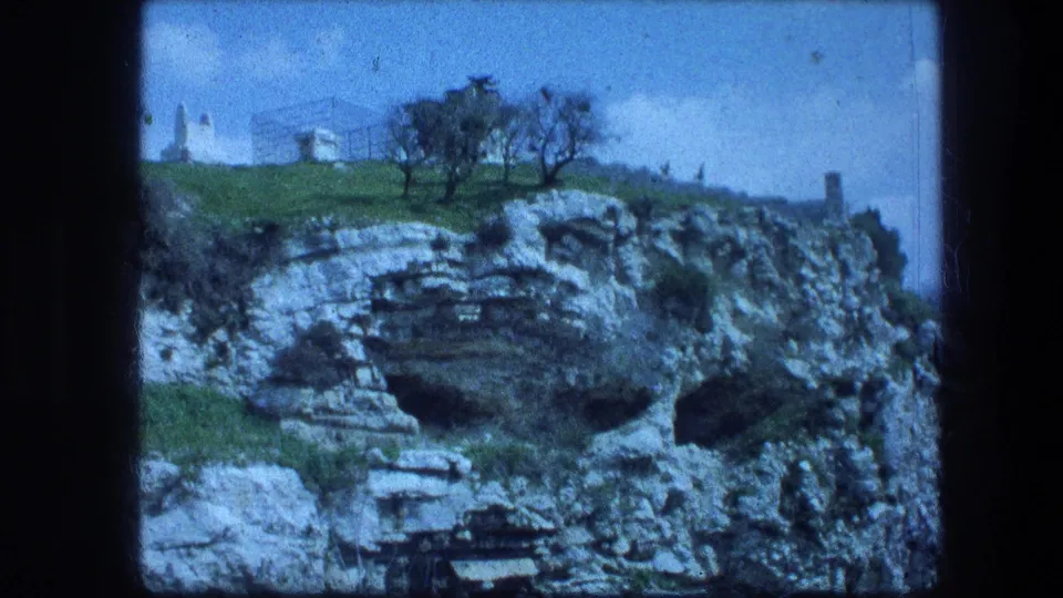 1976 8mm view of the Garden Tomb landscape, with a pale structure above a rocky cliff and trees on the ridge.