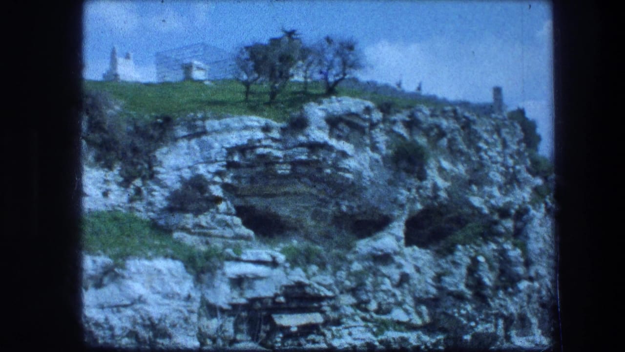Rocky limestone ledge with cave openings, grass, trees, and a small white building above.