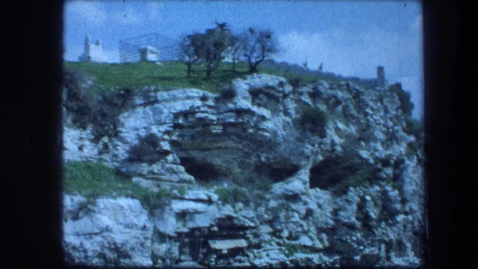 1976 8mm view of the Garden Tomb area in Jerusalem, showing a rocky cliff, cave openings, grassy ledge, trees, and white hilltop building.