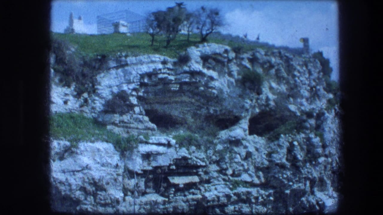 Layered rock face with dark cave openings below a grassy slope and small white building.