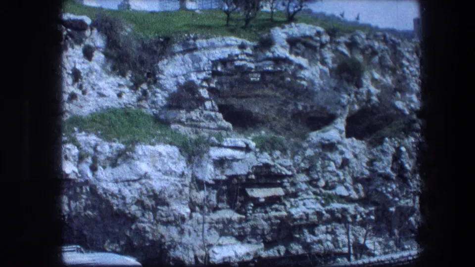 1976 8mm Garden Tomb view in Jerusalem, holding on a rocky ledge with cave openings, grass, sparse trees, and a blue sky.