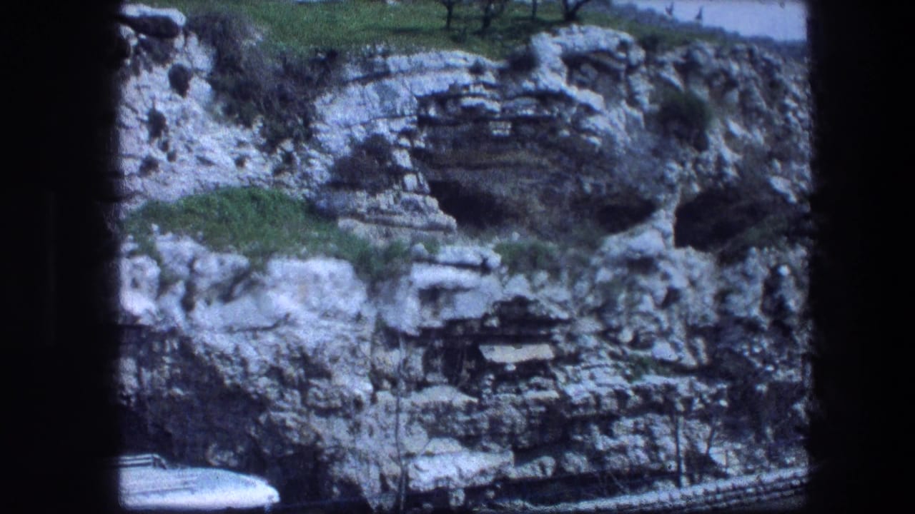 Layered rocky hillside with cave openings, grass patches, and a white vehicle at lower left.