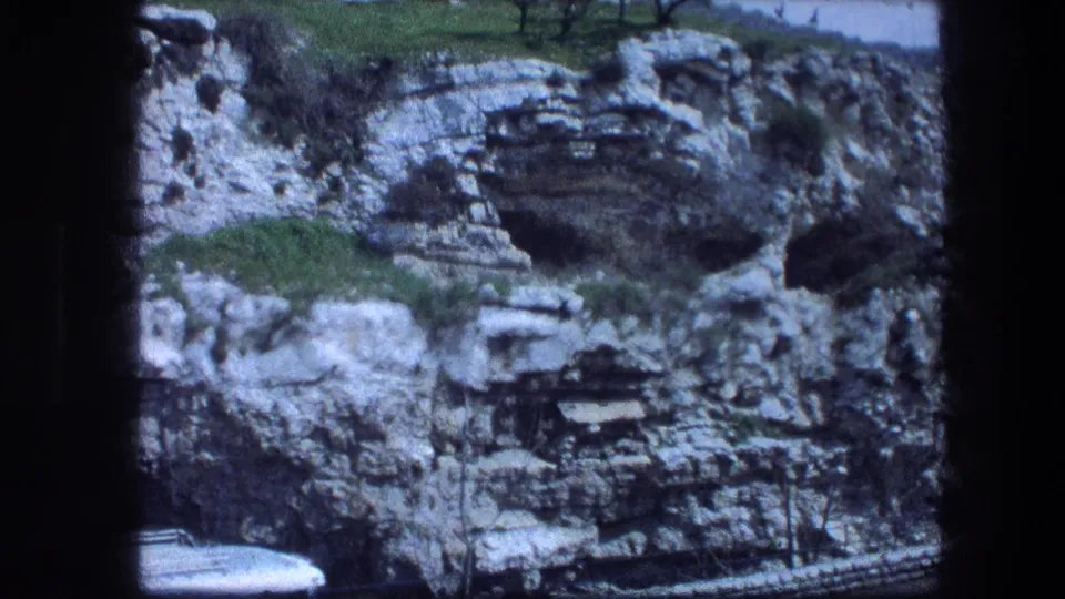 1976 8mm view of the Garden Tomb hillside in Jerusalem, showing limestone layers, dark caves, grass, and a white vehicle below.