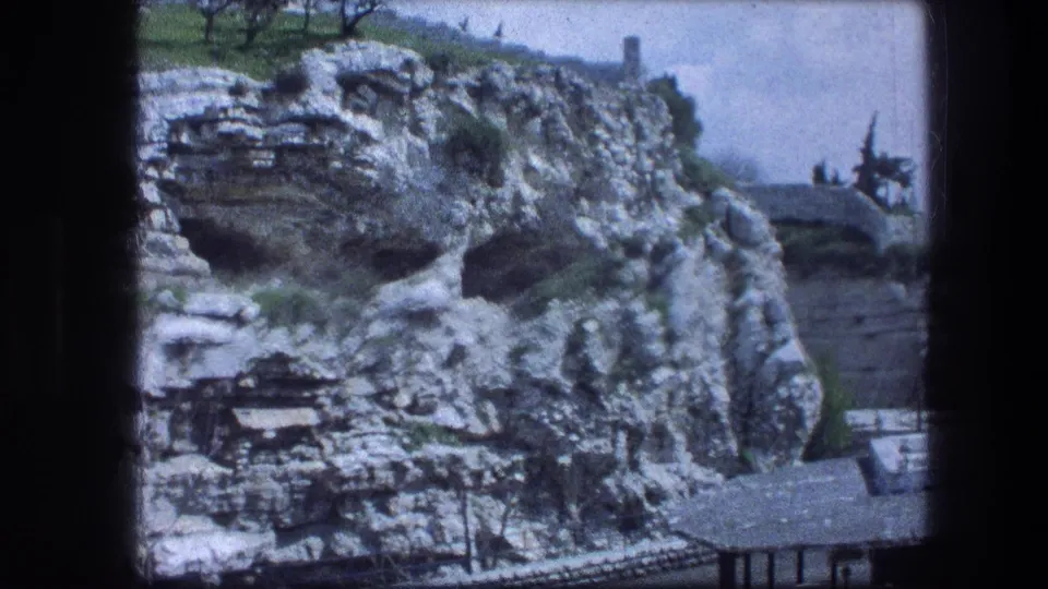 1976 Garden Tomb hillside view with layered rock, dark openings, grass at the top, and low built edges visible near the bottom.