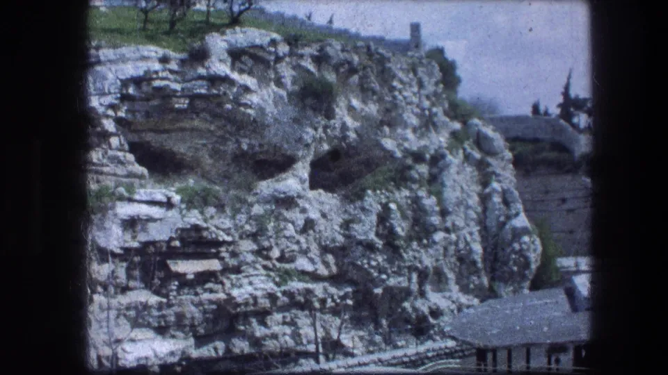Final 1976 frame of the Garden Tomb hillside in Jerusalem, with cave openings, green patches, pale sky, and a lower roofline.
