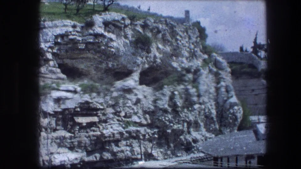 1976 8mm view of the Garden Tomb area in Jerusalem, showing pale limestone cliffs, shadowed openings, grass, and distant trees.