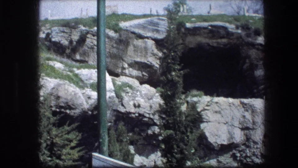 1976 visitor-shot frame of the Garden Tomb area, with a shadowed cave entrance, limestone ledges, shrubs, and a pole in the foreground.