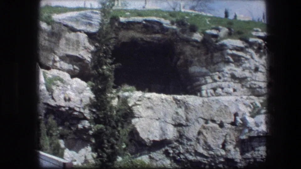 Garden Tomb rock face in Jerusalem, showing a broad shadowed opening, pale limestone, rough stonework, and small trees in faded 8mm color.