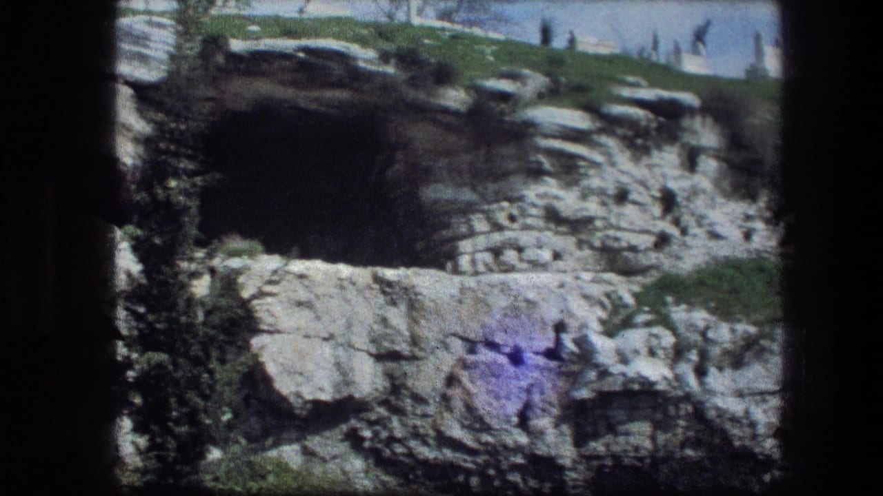 Layered limestone wall with dark cave opening, grass above, and 8mm frame edges.