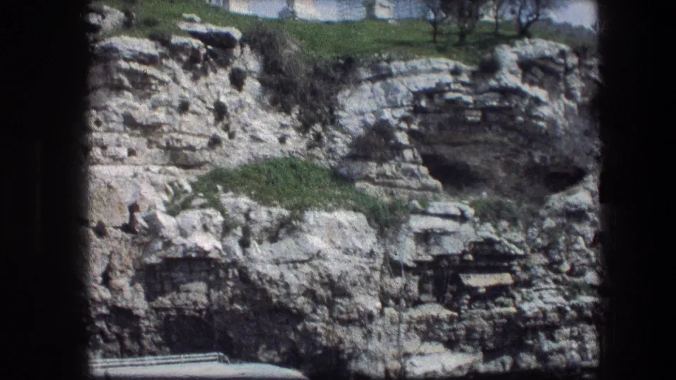 1976 home-movie frame of the Garden Tomb rock face, showing a large dark recess, layered stone, green vegetation, and a low ledge.