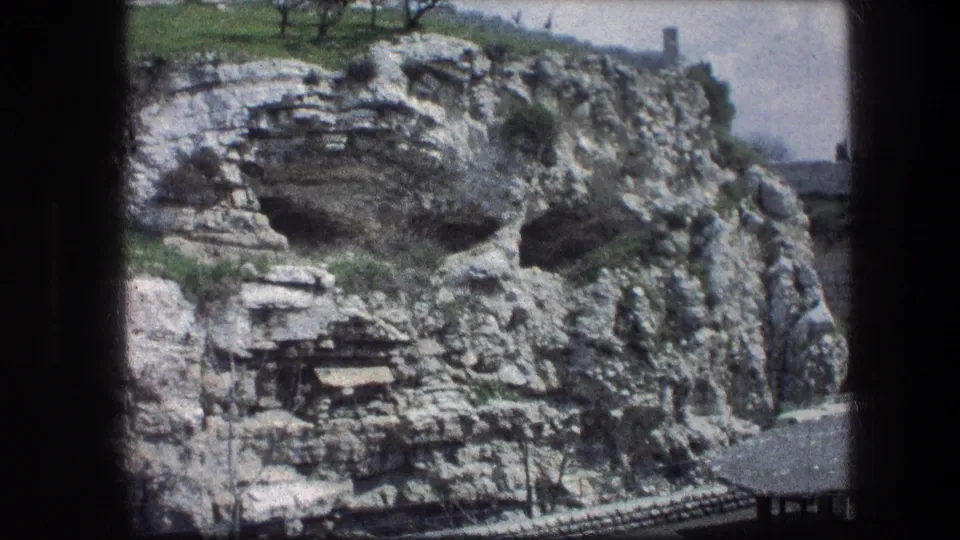 1976 8mm view of the Garden Tomb hillside in Jerusalem, showing rugged rock faces, dark openings, and a bit of greenery above.