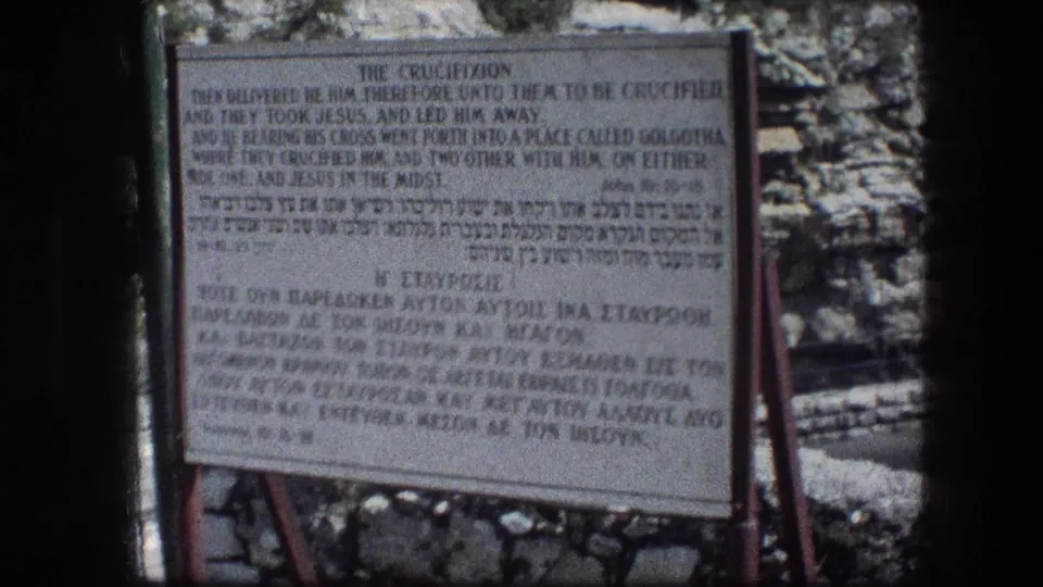 Close 8mm view of a Garden Tomb sign headed The Crucifixion, with English, Hebrew, and Greek text set against rocky Jerusalem terrain.