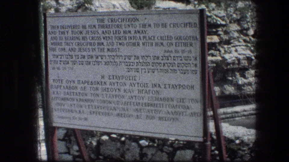 The Garden Tomb crucifixion sign fills the frame, with multilingual text, metal supports, and a rocky Jerusalem backdrop visible behind it.