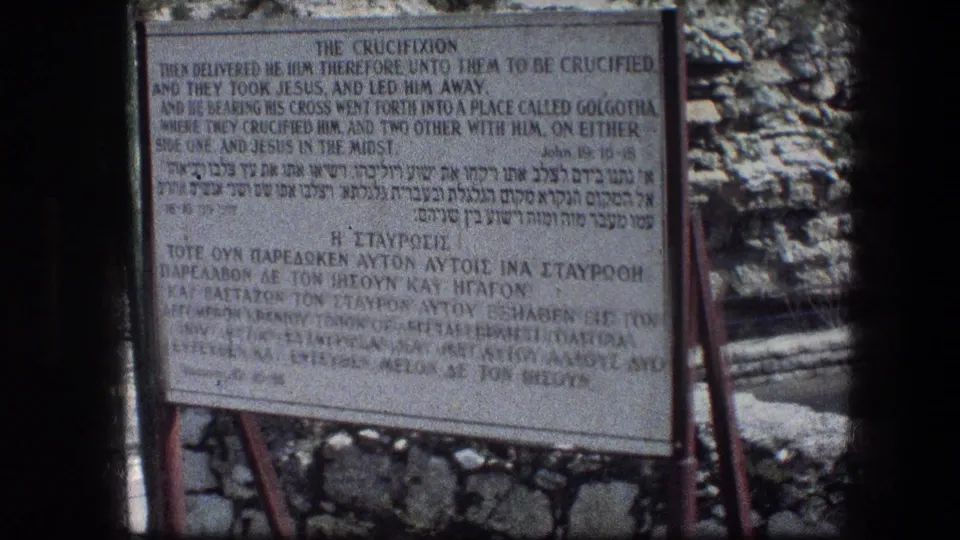 Soft, grainy 1976 8mm view of the Garden Tomb crucifixion sign, with multilingual text and stonework behind it.