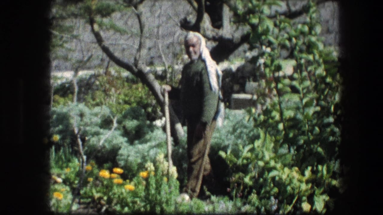 Elderly man with white head covering standing among garden plants.
