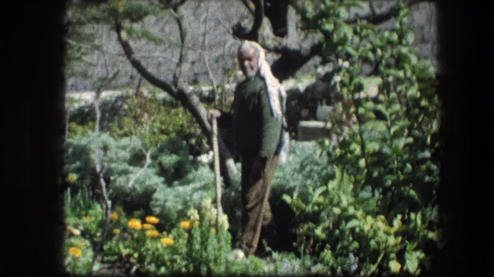 1976 Garden Tomb frame showing an elderly man in a green shirt standing in a lush garden with branches and flowers around him.