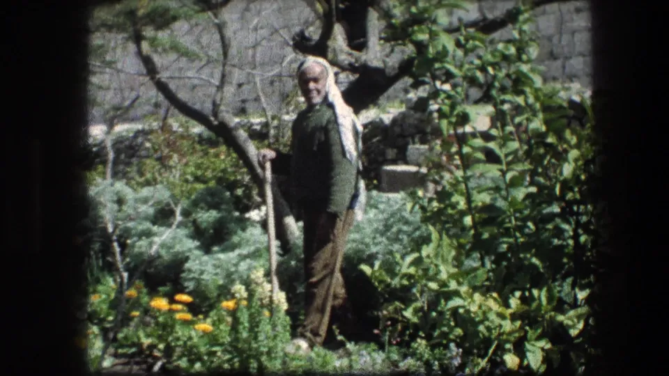 1976 8mm frame from the Garden Tomb area showing an elderly man with long white hair or head covering standing amid shrubs and flowers.