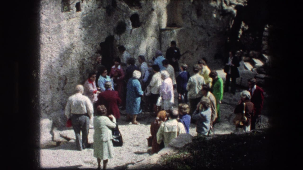 Small crowd standing near grey stone walls in bright sun.