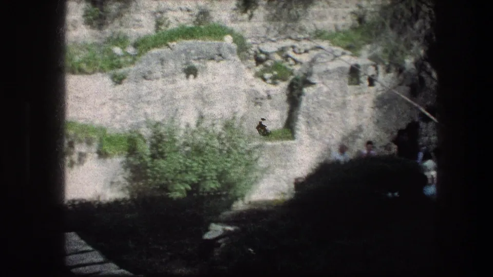 A wider 8mm frame shows the Garden Tomb area with pale rock surfaces, greenery, shadow, and tourists partly visible below.