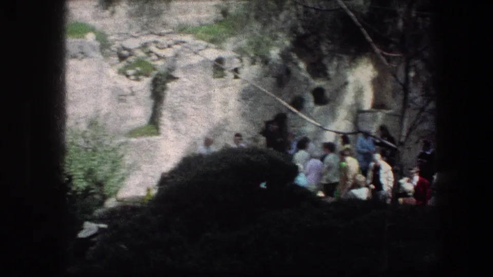 Tourists move along a Garden Tomb path in Jerusalem, framed by pale rock, plants, and strong shadow from the handheld 8mm camera.