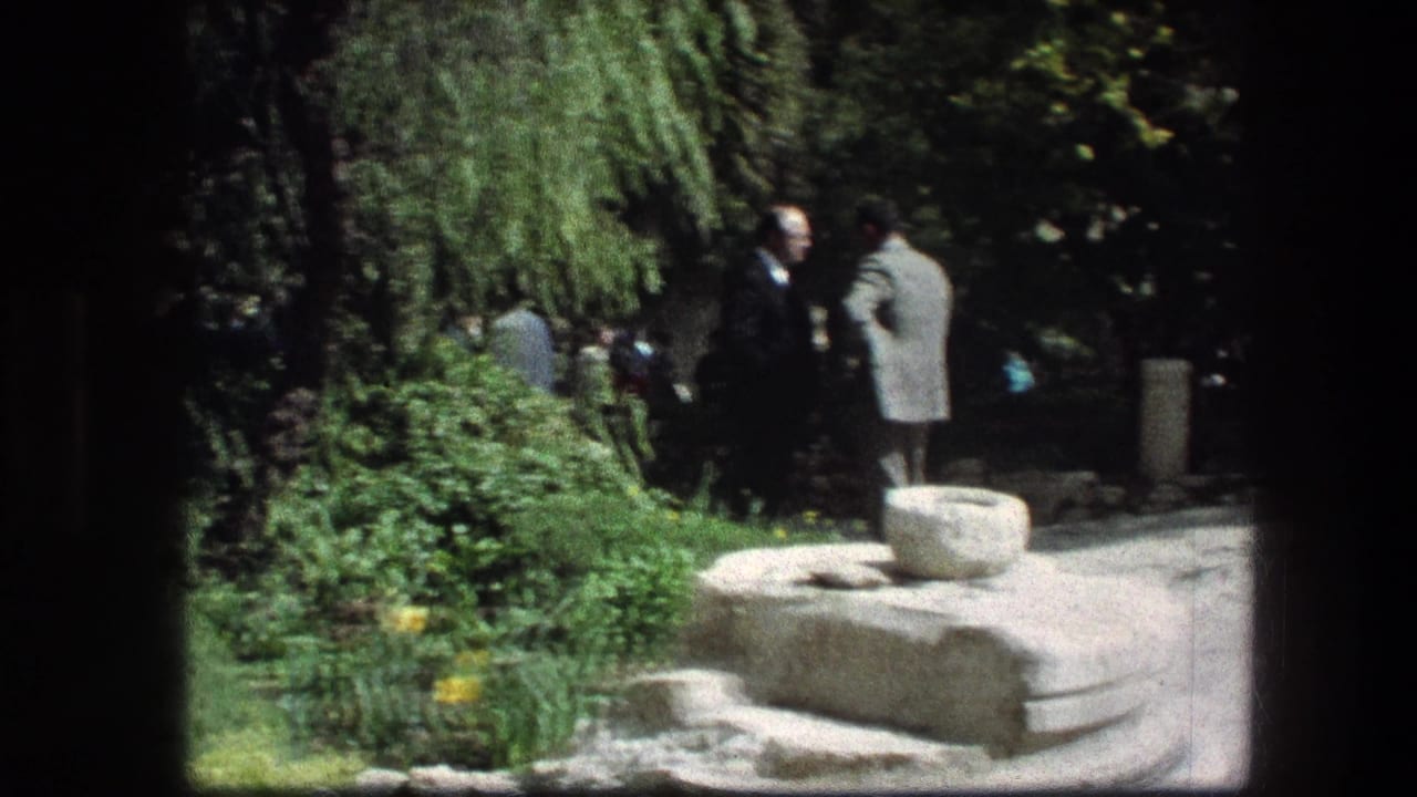 Two men stand talking beside a stone feature in a sunny garden path.
