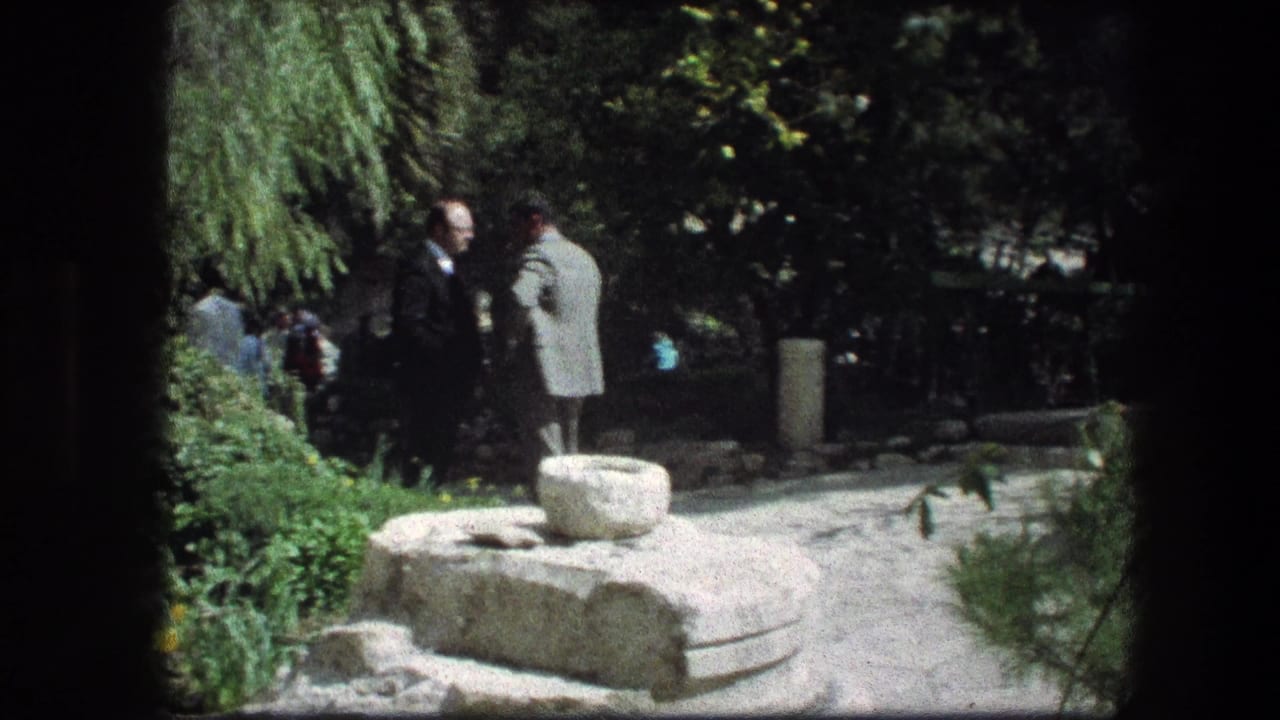 Two men stand in conversation near a stone feature at the Garden Tomb, with shaded greenery and other visitors visible behind them.