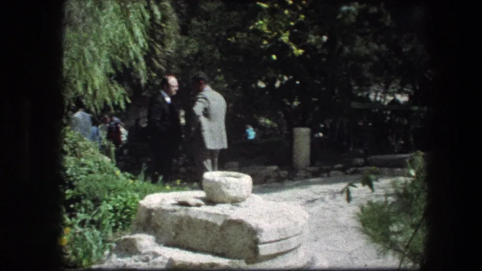 Two men stand in conversation near a stone feature at the Garden Tomb, with shaded greenery and other visitors visible behind them.