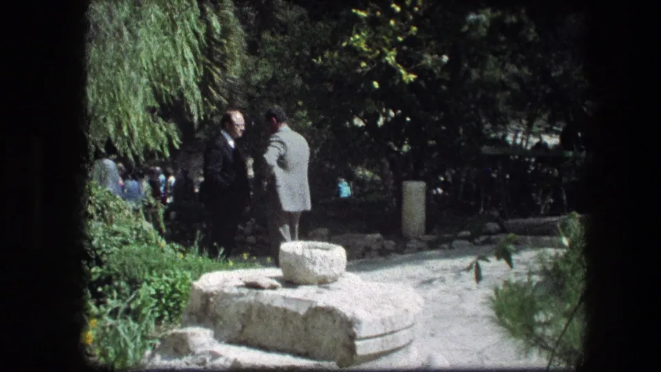 The 1976 Garden Tomb footage shows two men conversing near a stone feature, framed by shrubs, flowers, and deep tree shade.