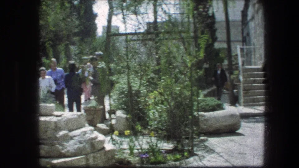 Tourists walk through the Garden Tomb grounds in Jerusalem, passing stone paths, garden beds, tall plants, and steps in 1976 home-movie footage.