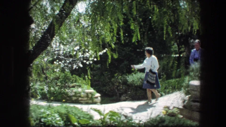 At the Garden Tomb in Jerusalem, the woman gestures while walking along a sunlit path beside shrubs and stone edging.