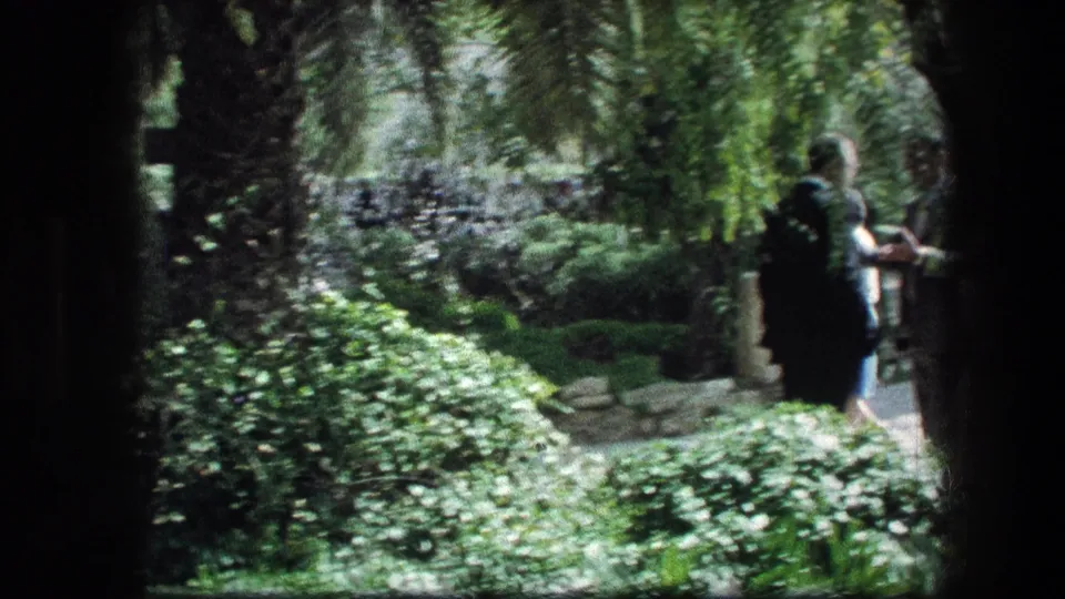 1976 8mm frame from the Garden Tomb area showing a woman near a shaded path with stone edging and thick plantings.