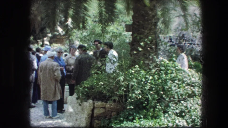 1976 home-movie frame of adult visitors standing and talking in the Garden Tomb garden in Jerusalem.