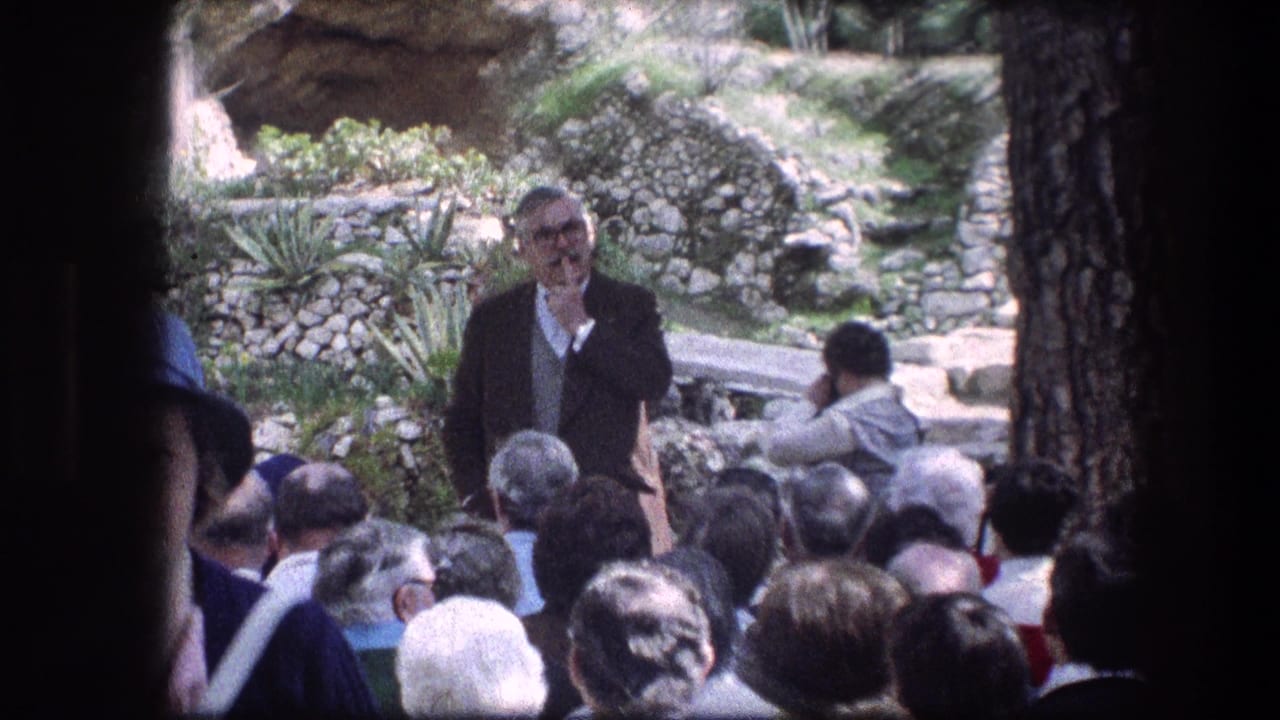 Man in grey jacket speaking to a seated group outdoors near stone garden walls.