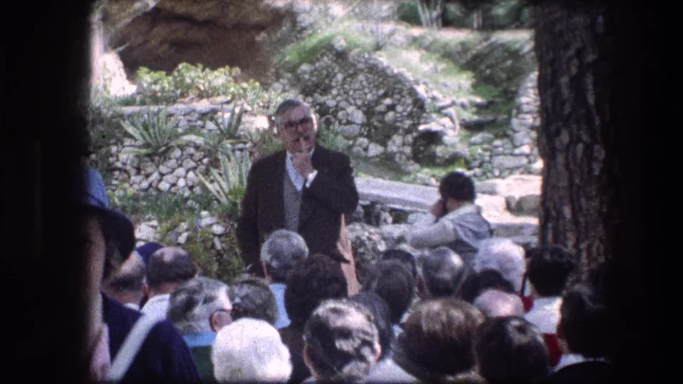 1976 8mm frame at the Garden Tomb in Jerusalem showing a man in a grey jacket addressing visitors outdoors.