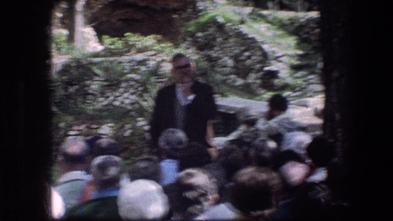 Soft-focus view of a man speaking before a seated crowd in a garden setting.