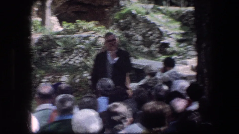 Blurred 1976 Garden Tomb frame with a speaker facing visitors among stonework, trees, and planted areas.