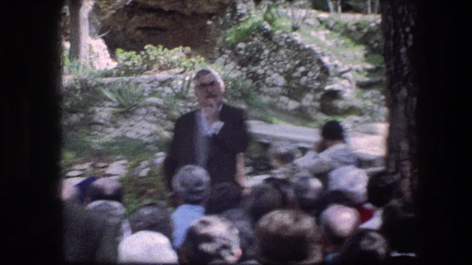 1976 Garden Tomb frame showing the speaker and audience outdoors, with low stone walls and vegetation behind them.