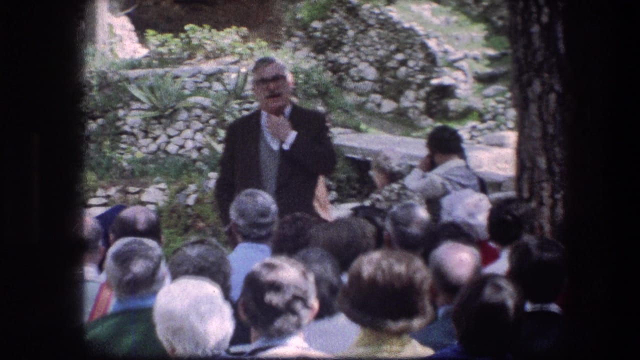 Audience seated outdoors while a man in a dark suit speaks near stone garden walls.