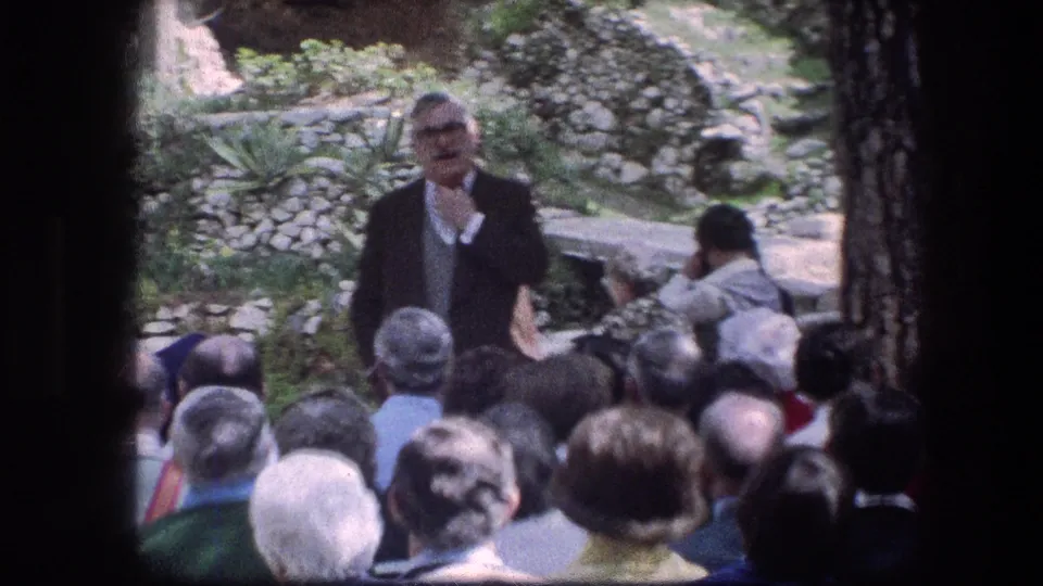 1976 8mm frame at the Garden Tomb in Jerusalem showing adults seated outdoors as a suited man addresses them near stone walls and greenery.